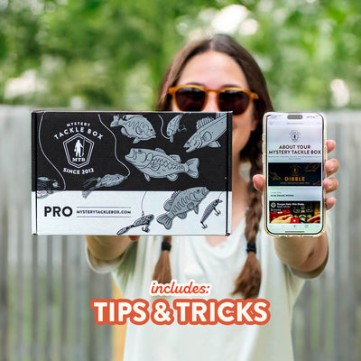 Person holding a fishing tackle box and phone with text 'Mystery Tackle Box' and 'includes: Tips & Tricks'.